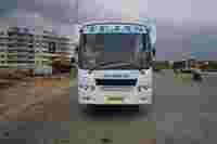 A white bus labeled "TEJAS" and "SRI MARUTHI" is parked on a road near residential buildings under a cloudy sky.