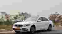 A white luxury sedan drives on a road, surrounded by flowering trees and a clear sky.