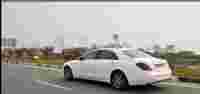 A white luxury sedan drives along a highway lined with flowering plants and signage in the background.
