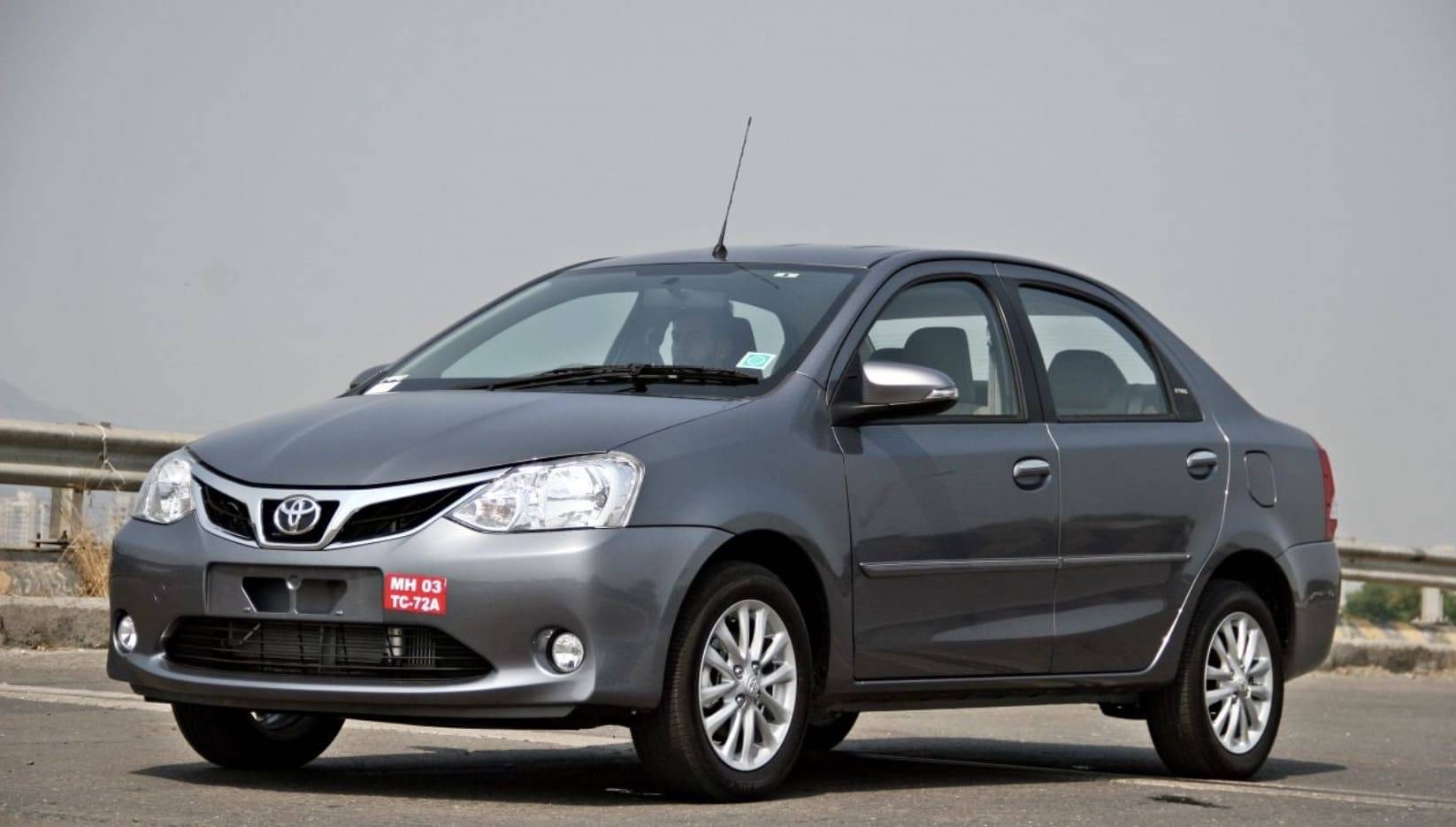 A gray Toyota sedan parked on a highway, with a driver visible in the front seat and a blurred cityscape in the background.