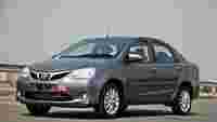 A gray Toyota sedan parked on a highway, with a driver visible in the front seat and a blurred cityscape in the background.