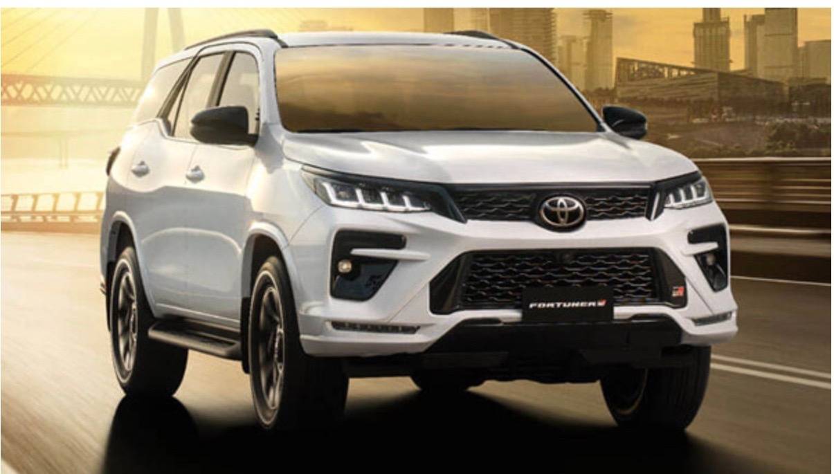 White Toyota Fortuner SUV driving on a road with a city skyline and sunset in the background.