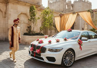 A smiling man in traditional attire walks beside a decorated white luxury car, set against an elegant outdoor backdrop.