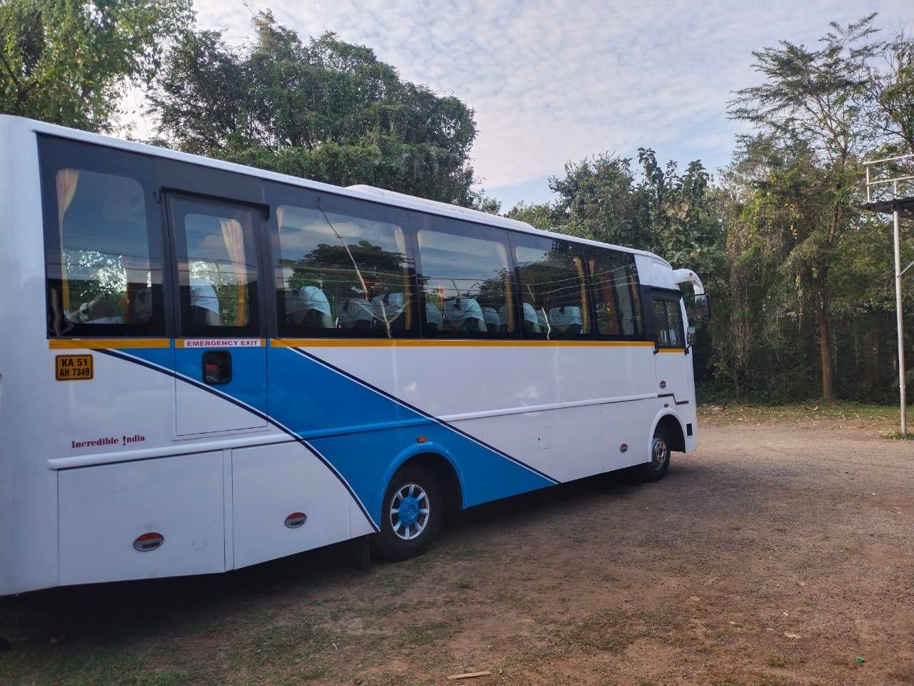 A white and blue bus is parked on a gravel surface, surrounded by green trees and shrubbery under a cloudy sky.