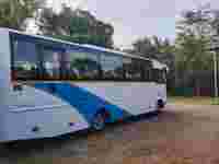 A white and blue bus is parked on a gravel surface, surrounded by green trees and shrubbery under a cloudy sky.