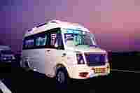 A white bus labeled "TEJAS" is parked on a highway at dusk, with illuminated interiors and another bus in the background.