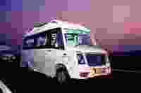 A white bus labeled "TEJAS" is parked on a highway at dusk, with illuminated interiors and another bus in the background.