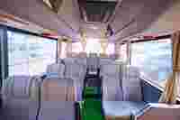 Interior of a spacious, empty bus with beige seats, green carpet, and sunlight streaming through the curtains.