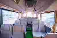 Interior of a spacious, empty bus with beige seats, green carpet, and sunlight streaming through the curtains.