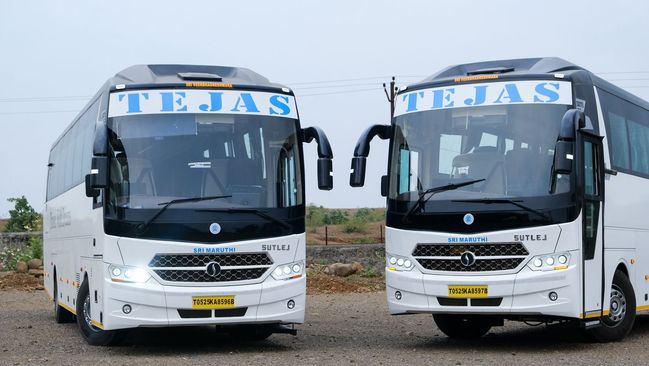 Two white buses labeled "TEJAS" parked on gravel, featuring sleek designs and minimalist branding, set against a muted backdrop.