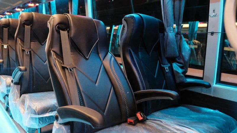 Luxurious black bus seats with armrests, partially covered in plastic, lined up against large windows in a modern setting.