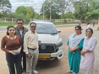 Five people, two men and three women, stand near a white Toyota vehicle on a roadside, surrounded by greenery.