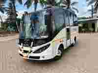 A white and black bus adorned with floral garlands parked on a paved area, surrounded by palm trees and a blue sky.