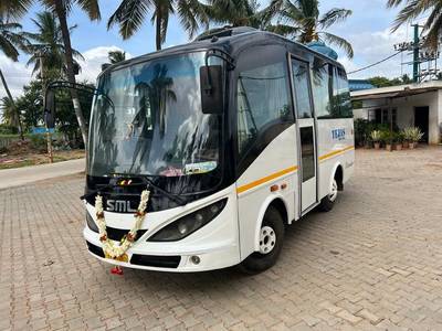 A white and black SML bus adorned with floral garlands parked on a paved area, surrounded by palm trees and blue sky.