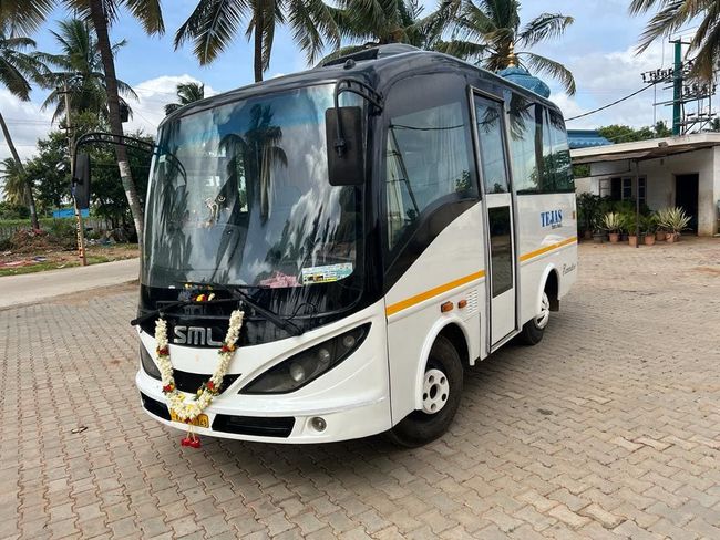 A white minibus with a floral garland parked on a tiled surface, surrounded by palm trees and a building in the background.