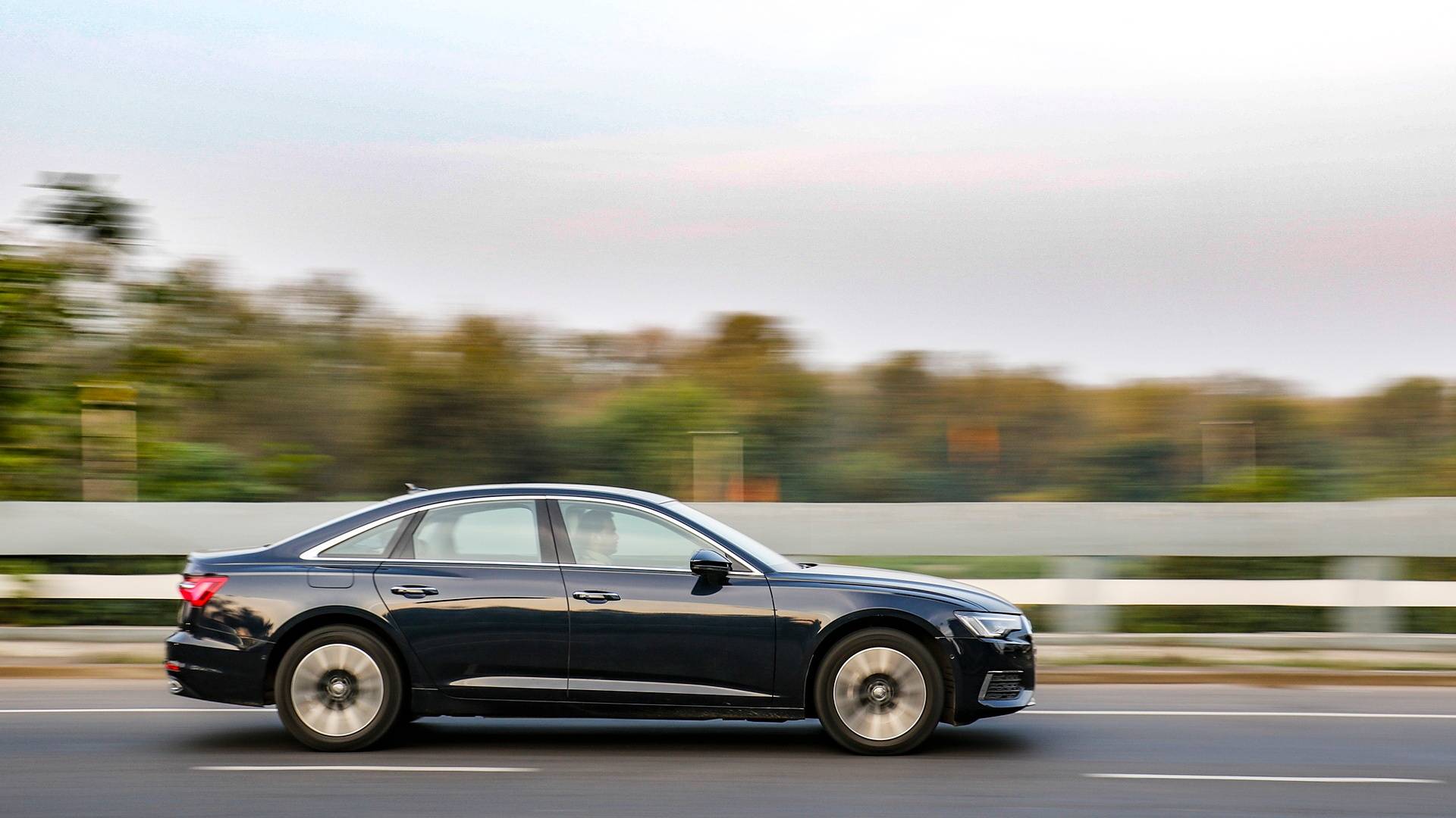 A black sedan drives quickly on a highway, with blurred greenery in the background suggesting high speed.