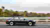 A black sedan drives quickly on a highway, with blurred greenery in the background suggesting high speed.