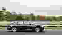 A black sedan drives quickly on a highway, with blurred greenery in the background suggesting high speed.