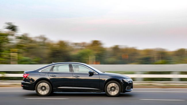 A black sedan drives quickly on a highway, with blurred greenery in the background suggesting high speed.