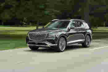 A sleek gray luxury SUV drives along a winding road, surrounded by green trees and an open landscape.
