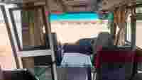 Interior of a bus showing the driver's seat, dashboard, and large windows overlooking a beach with blue water.