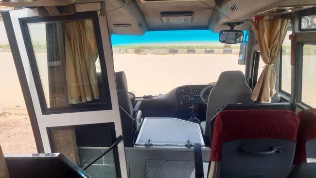 An empty bus interior featuring rows of blue seats with armrests and a red carpet, illuminated by natural light through windows.