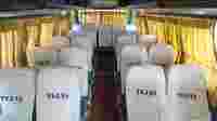 Interior of a bus featuring rows of white seats with "TEJAS" branding, warm lighting, and curtained windows.