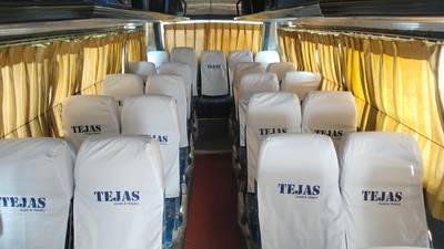 Interior of a bus with rows of covered seats labeled "TEJAS," illuminated by natural light from curtains.