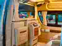 Luxurious van interior featuring beige leather seats, wooden accents, curtains, and a bright overhead light.