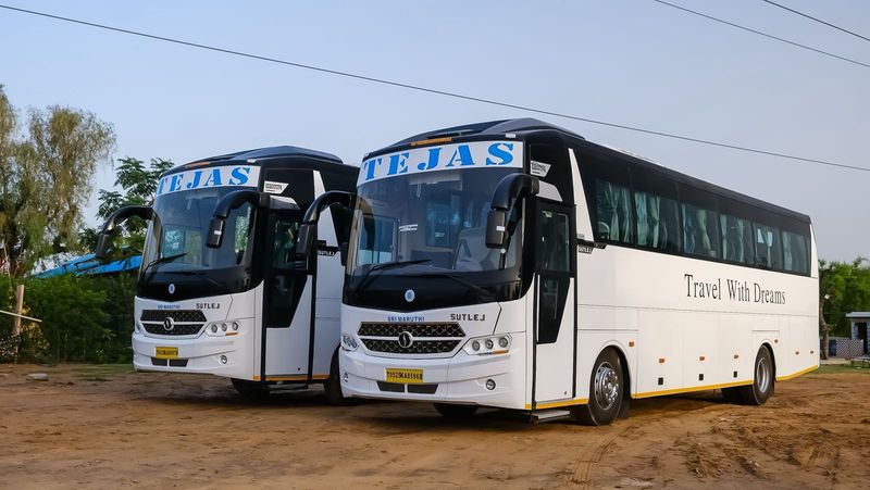 Two spacious, white tour buses parked on sandy ground, labeled "Tejas" and "Travel With Dreams." Green foliage in the background.