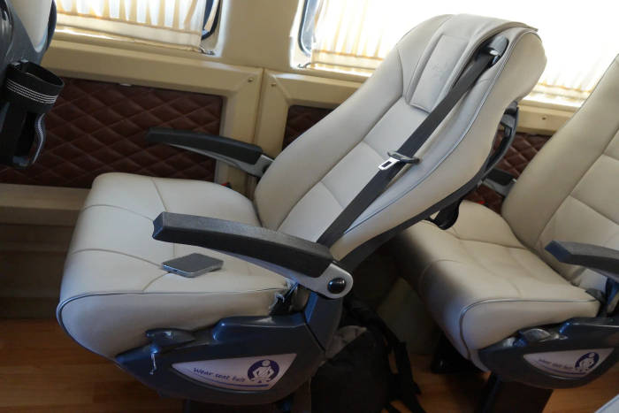A spacious, beige airplane seat with an adjustable armrest and a seatbelt, next to a backpack and patterned wall.