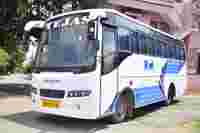 A white and blue bus with "TEJAS" on the front parked on a quiet street lined with trees and buildings.