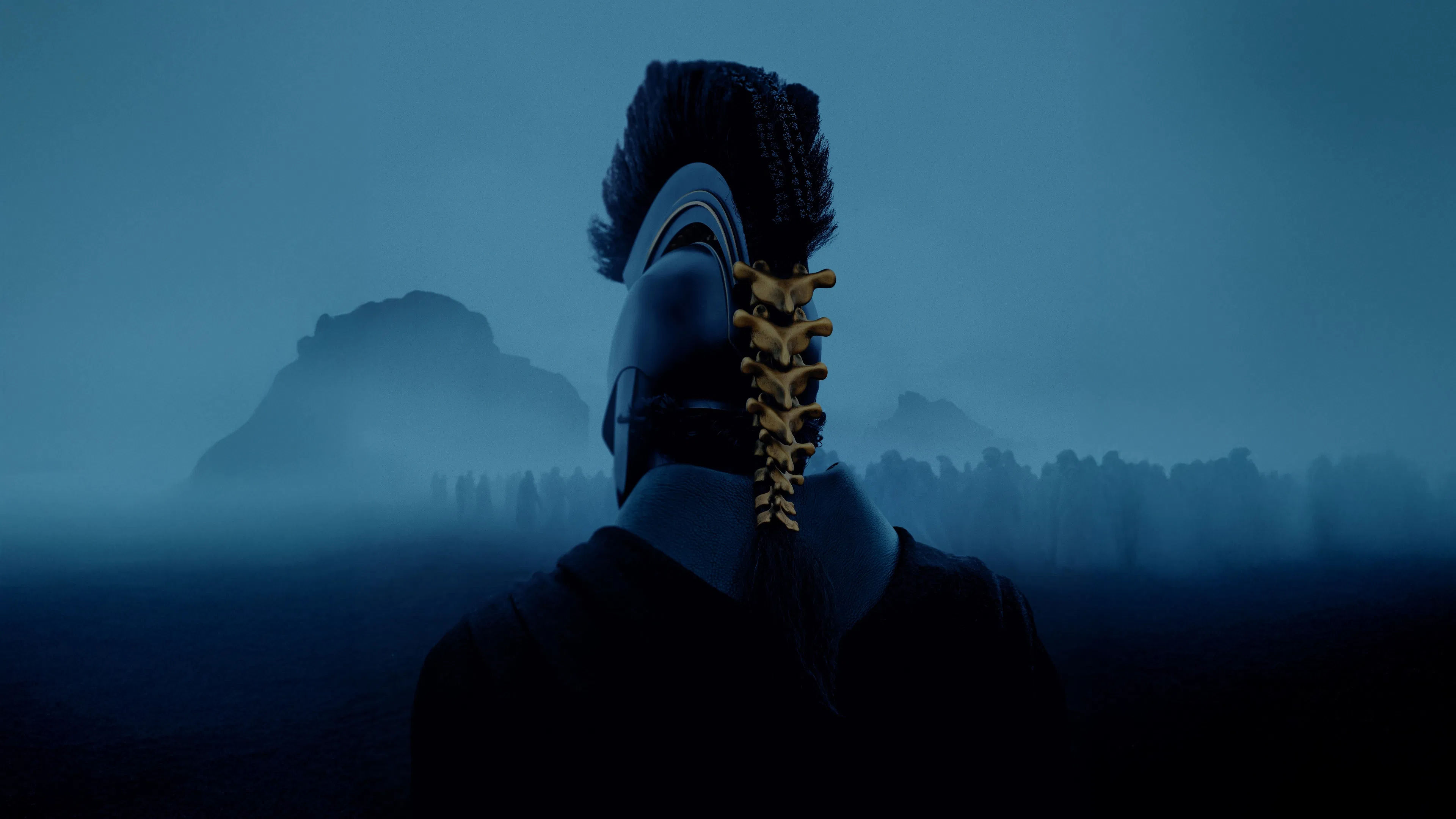 Back-facing view of a warrior wearing a black ancient helmet with a golden spine crest, looking out at a crowd of silhouettes in a misty, dark blue landscape.