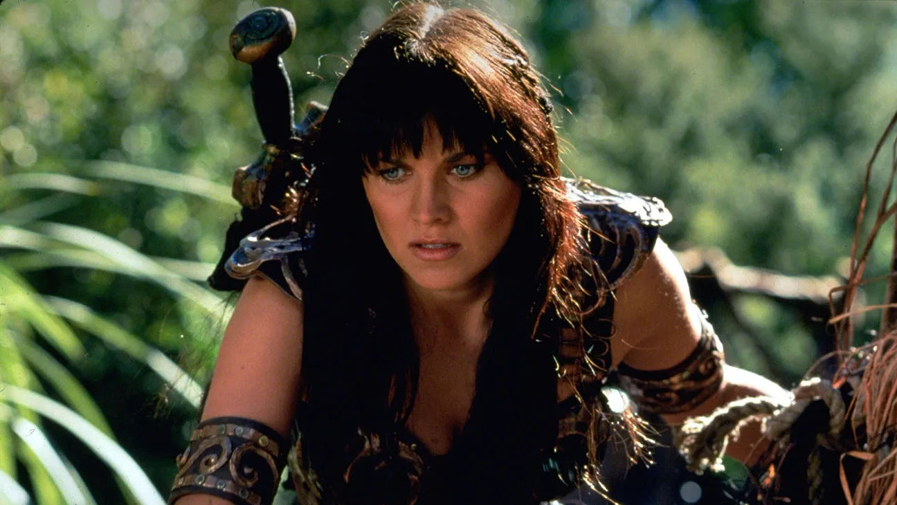 Close-up of dark haired woman, leaning forward with a focused expression, wearing dark leather armor and a sword strapped to her back.