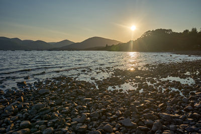 Sunset over Loch Lomond: Witness the perfect harmony of nature as the sun paints the sky in fiery hues while a serene coastal scene unfolds before your eyes.