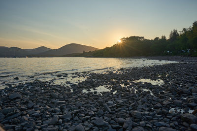 Sunset over Loch Lomond: Witness the perfect harmony of nature as the sun paints the sky in fiery hues while a serene coastal scene unfolds before your eyes.