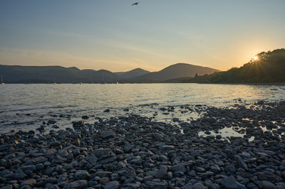 Sunset over Loch Lomond: Witness the perfect harmony of nature as the sun paints the sky in fiery hues while a serene coastal scene unfolds before your eyes.