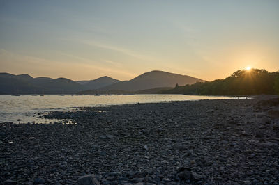 Sunset over Loch Lomond: Witness the perfect harmony of nature as the sun paints the sky in fiery hues while a serene coastal scene unfolds before your eyes.