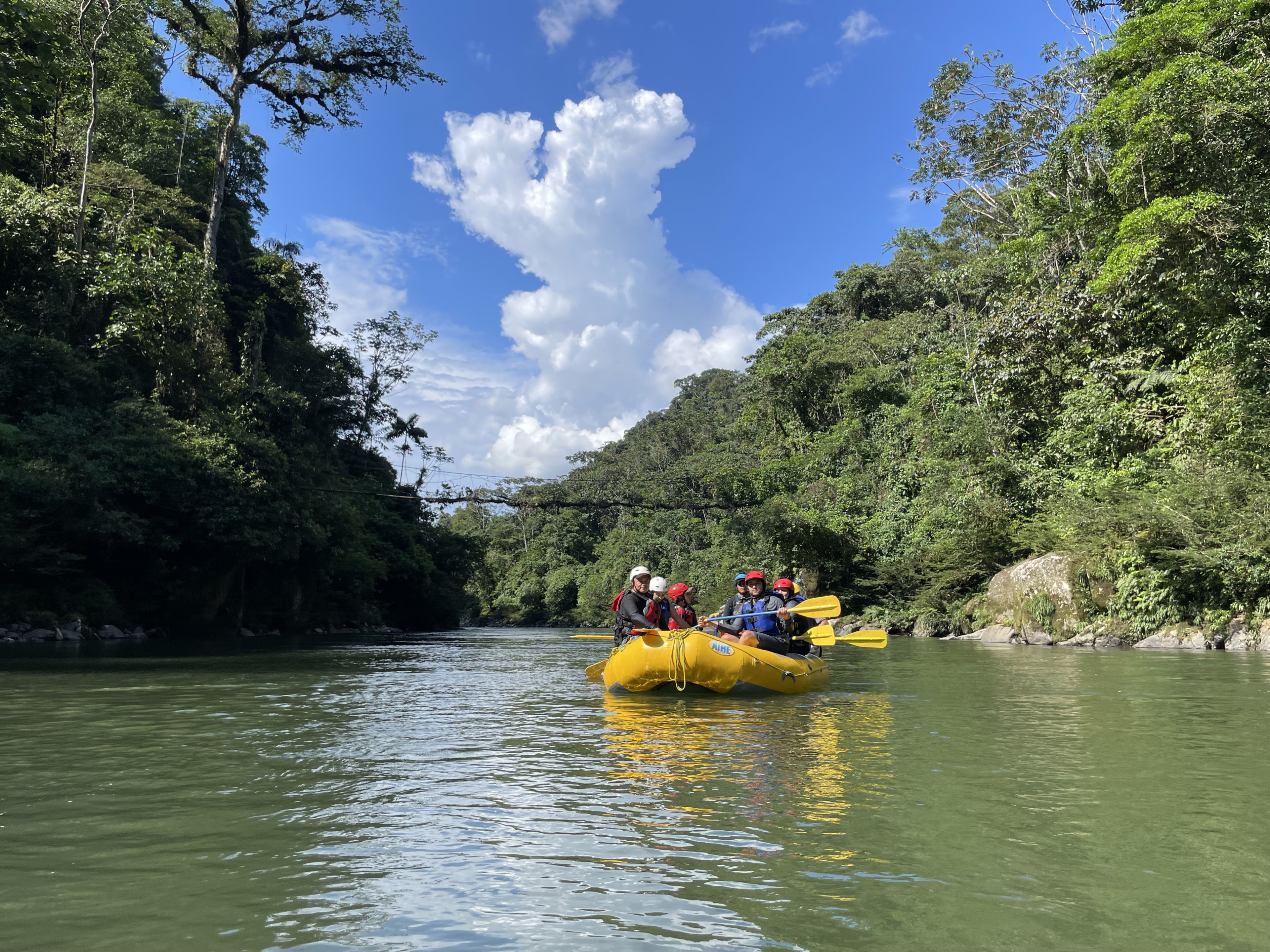 Group of people rafting on a river surrounded by lush greenery and blue sky.