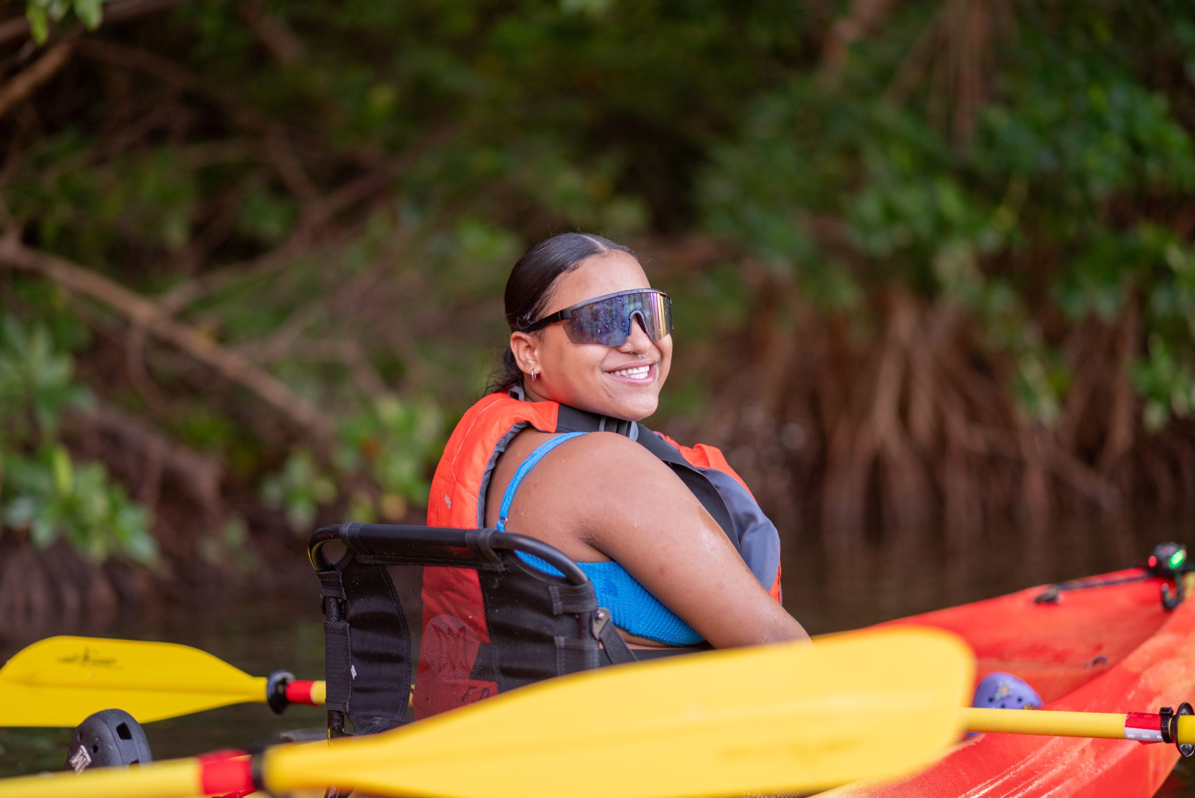 Smiling person in a kayak wearing sunglasses and a life jacket in a natural setting.