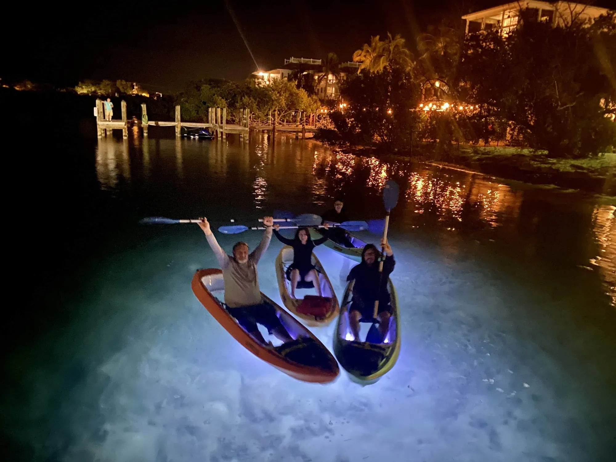 Four people kayaking at night with illuminated kayaks in calm water.
