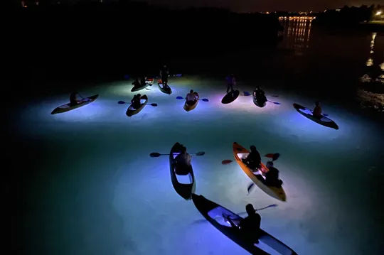 Kayakers illuminated by underwater lights on a dark waterway at night.
