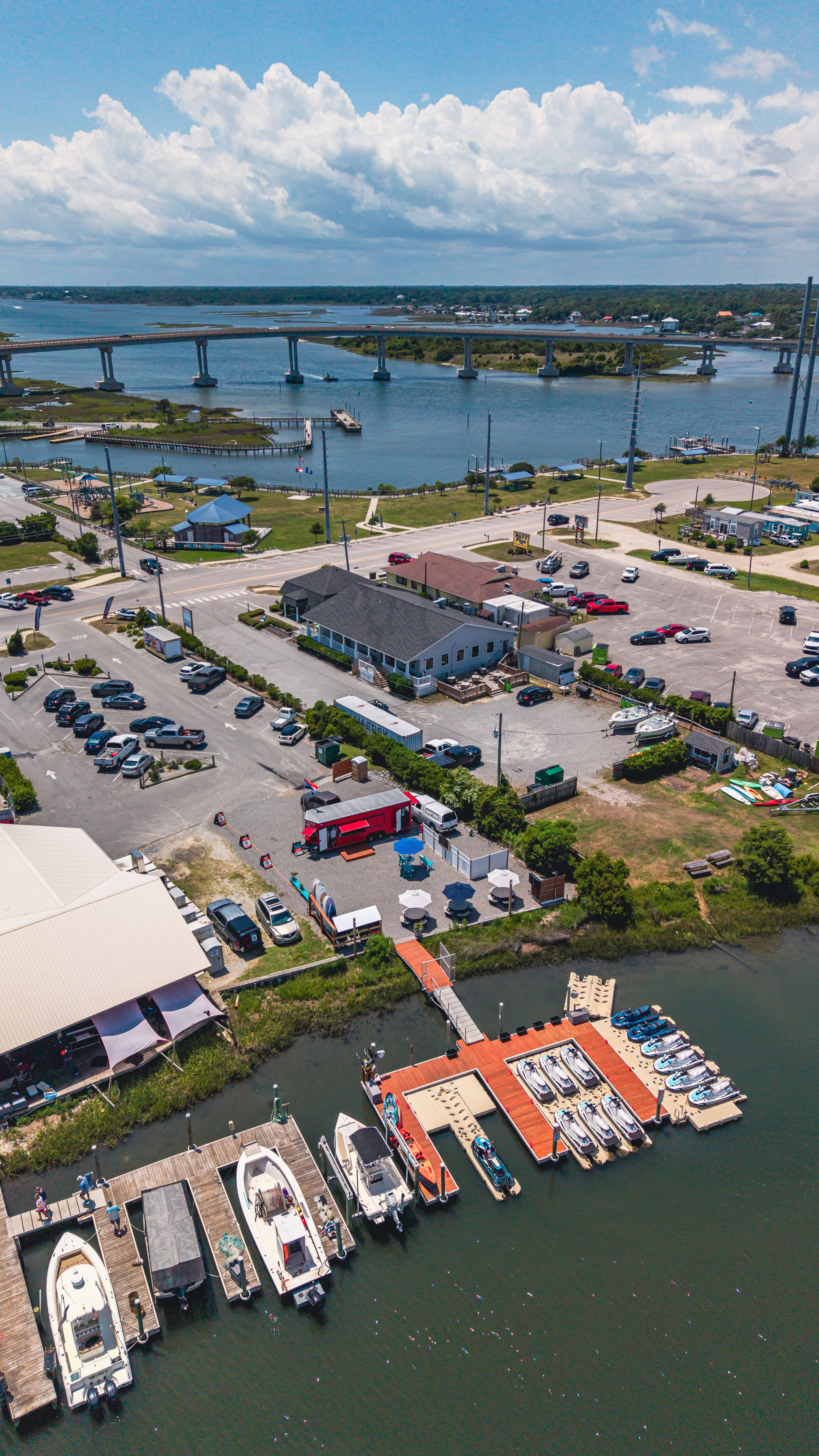 Aerial view of a marina with boats, parking, and a bridge in the background.