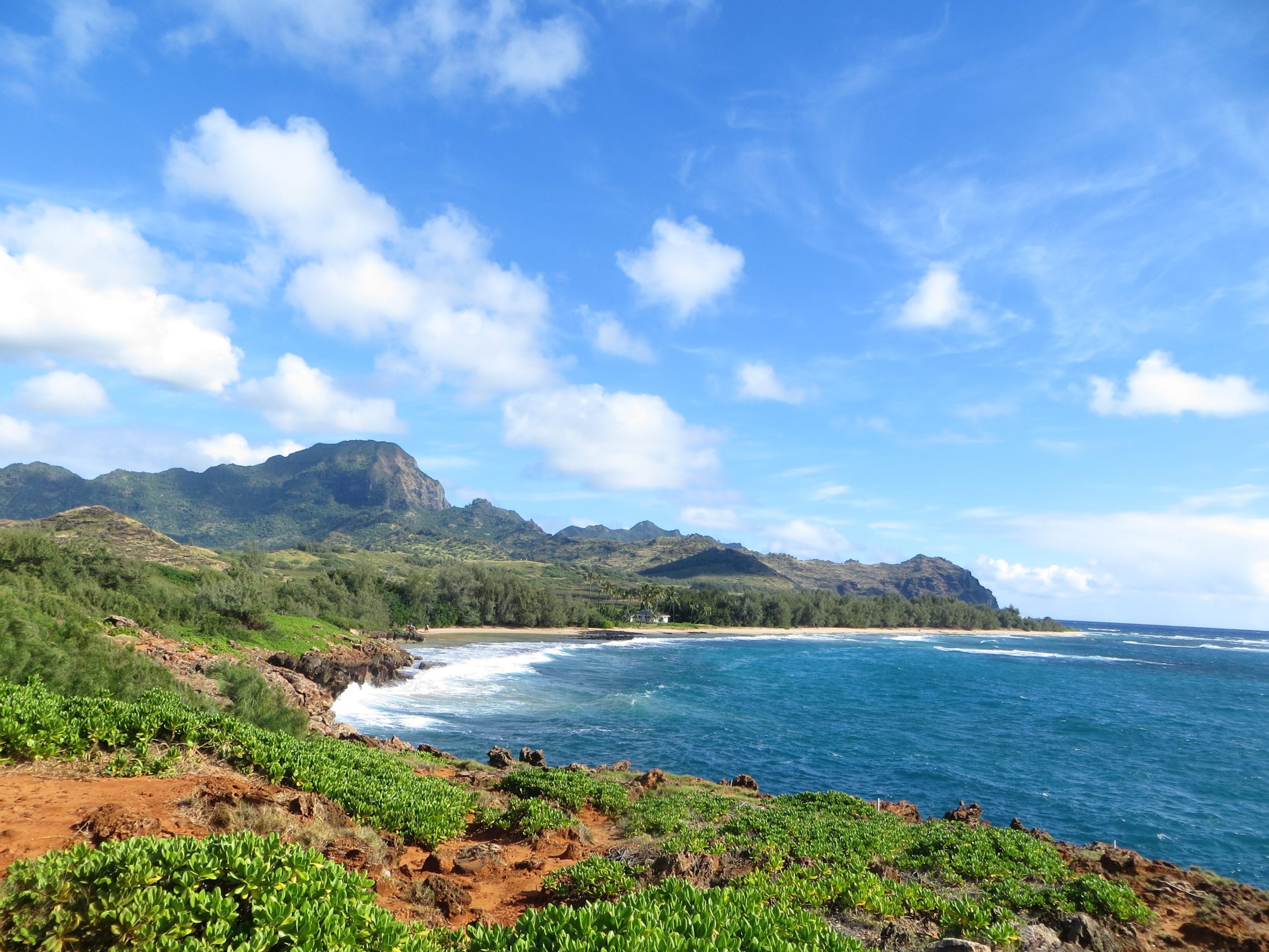Coastal view with mountains, blue water, and green vegetation under a partly cloudy sky.