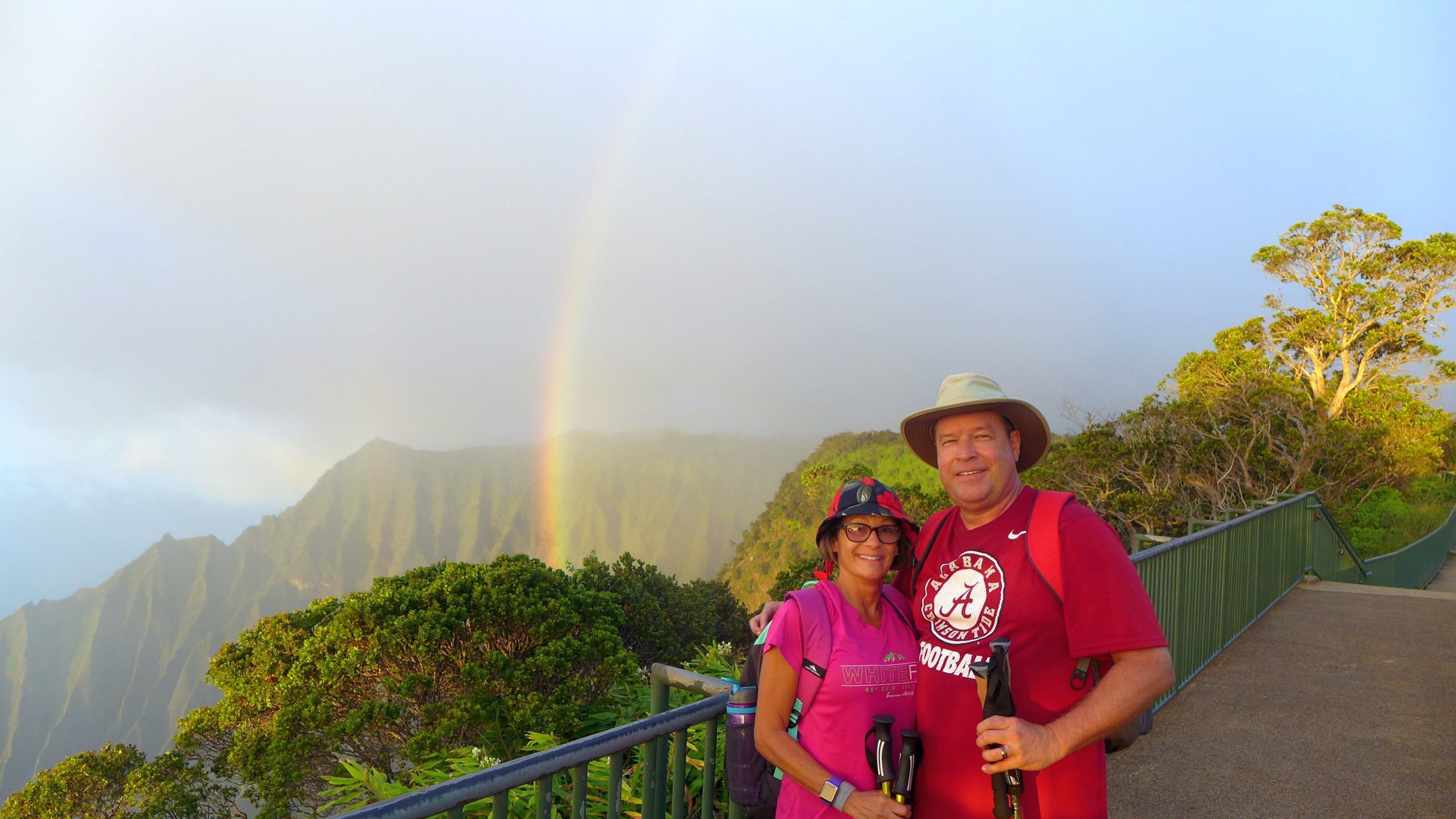 Couple posing with hiking gear and a rainbow in the background on a mountain trail.