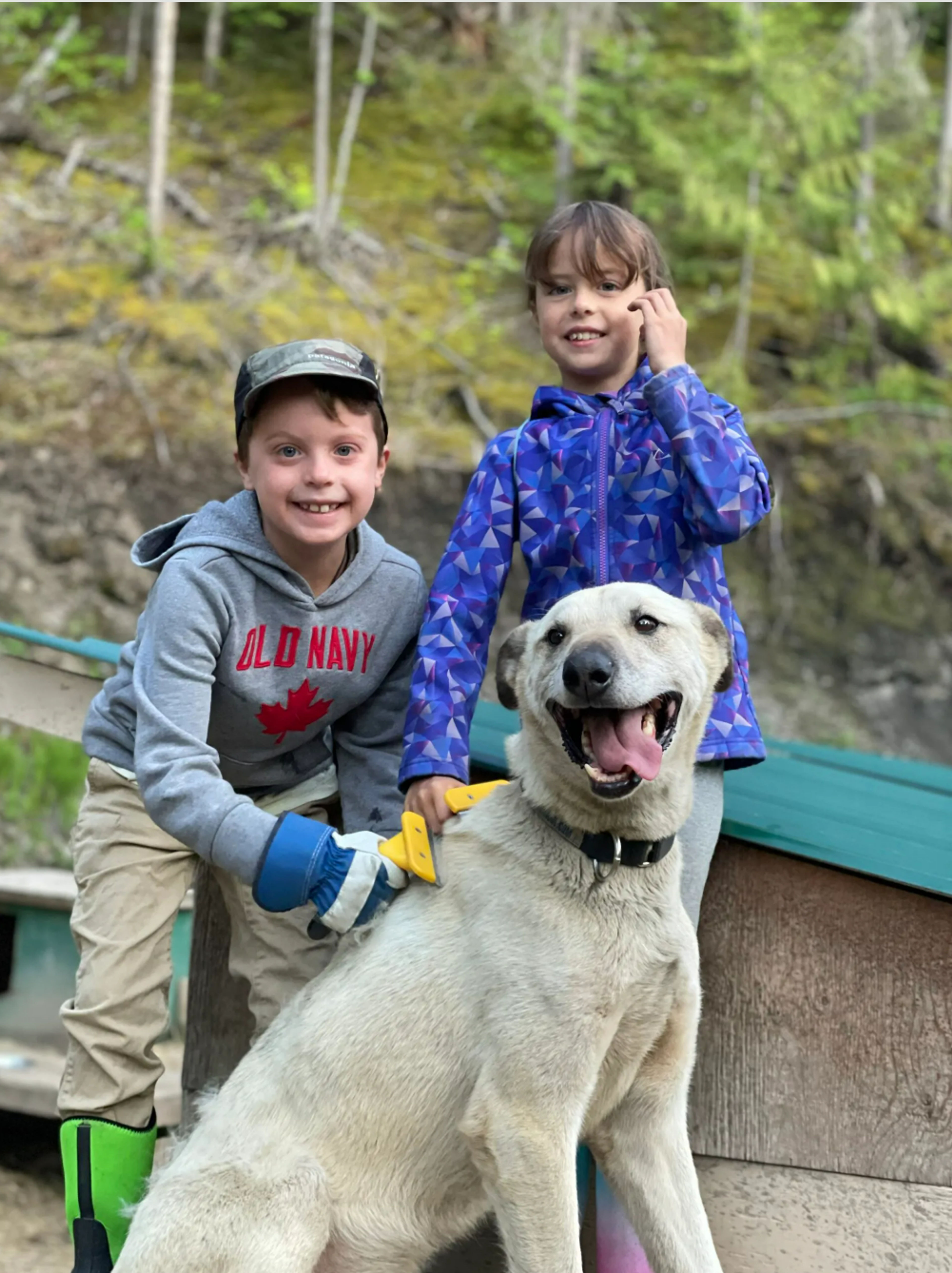 Two children smiling with a dog in a forested area