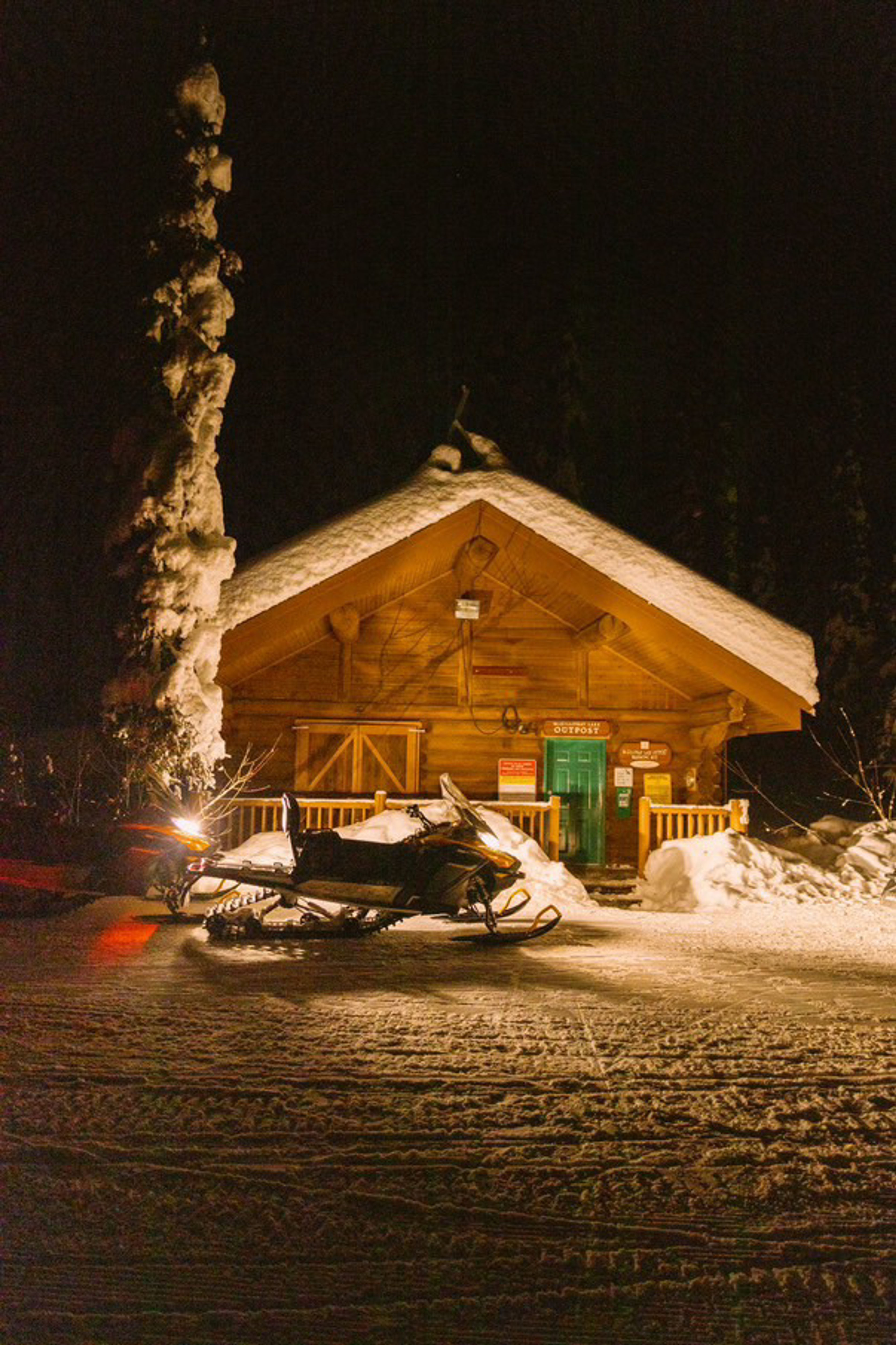 A cozy wooden cabin covered in snow at night with snowmobiles parked outside.