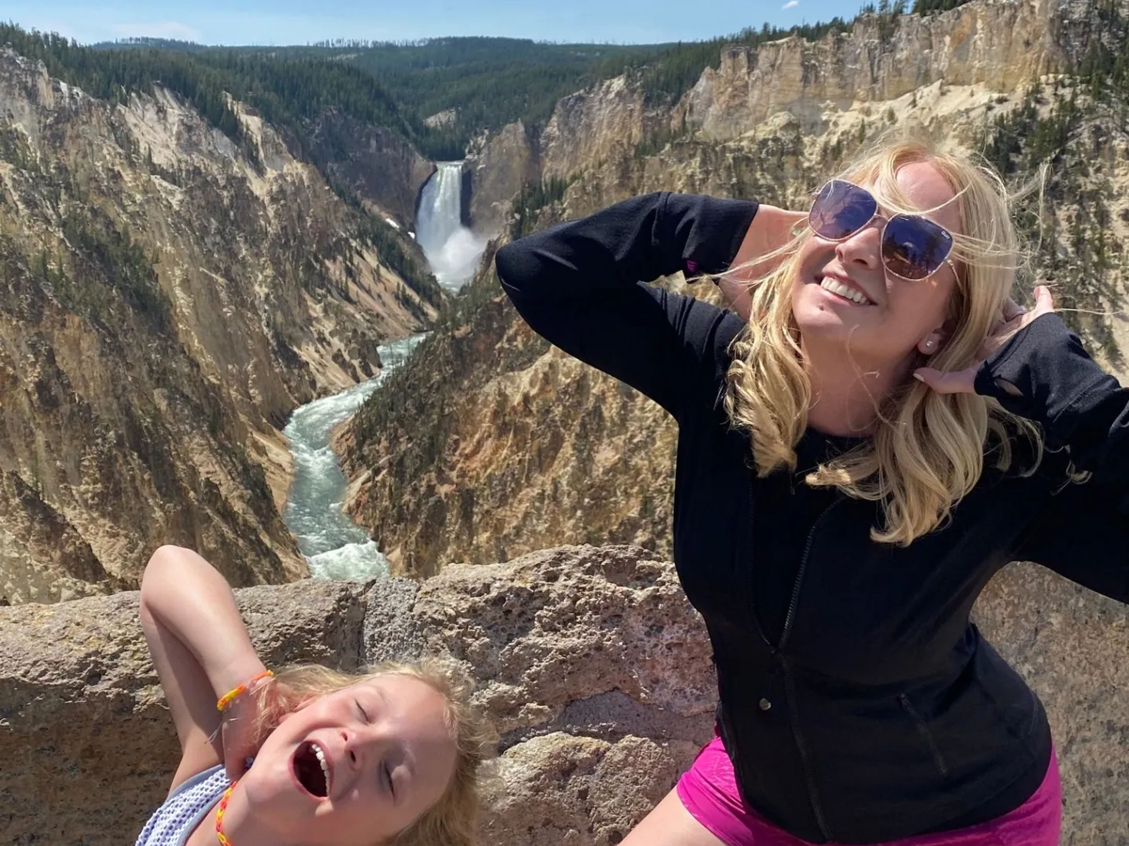 A woman and a girl joyfully pose at a scenic overlook with a waterfall in the background.