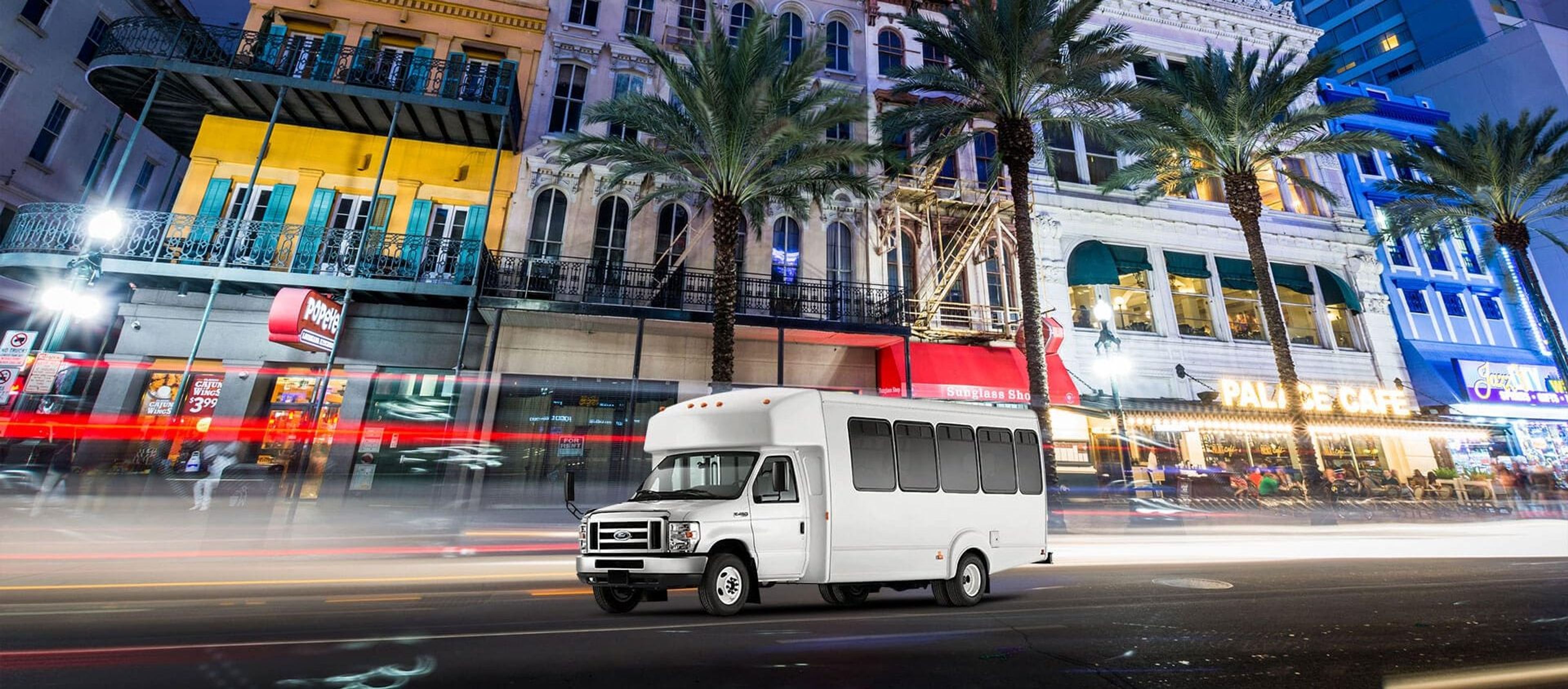 A white shuttle bus parked on a busy street with palm trees and colorful buildings at night.