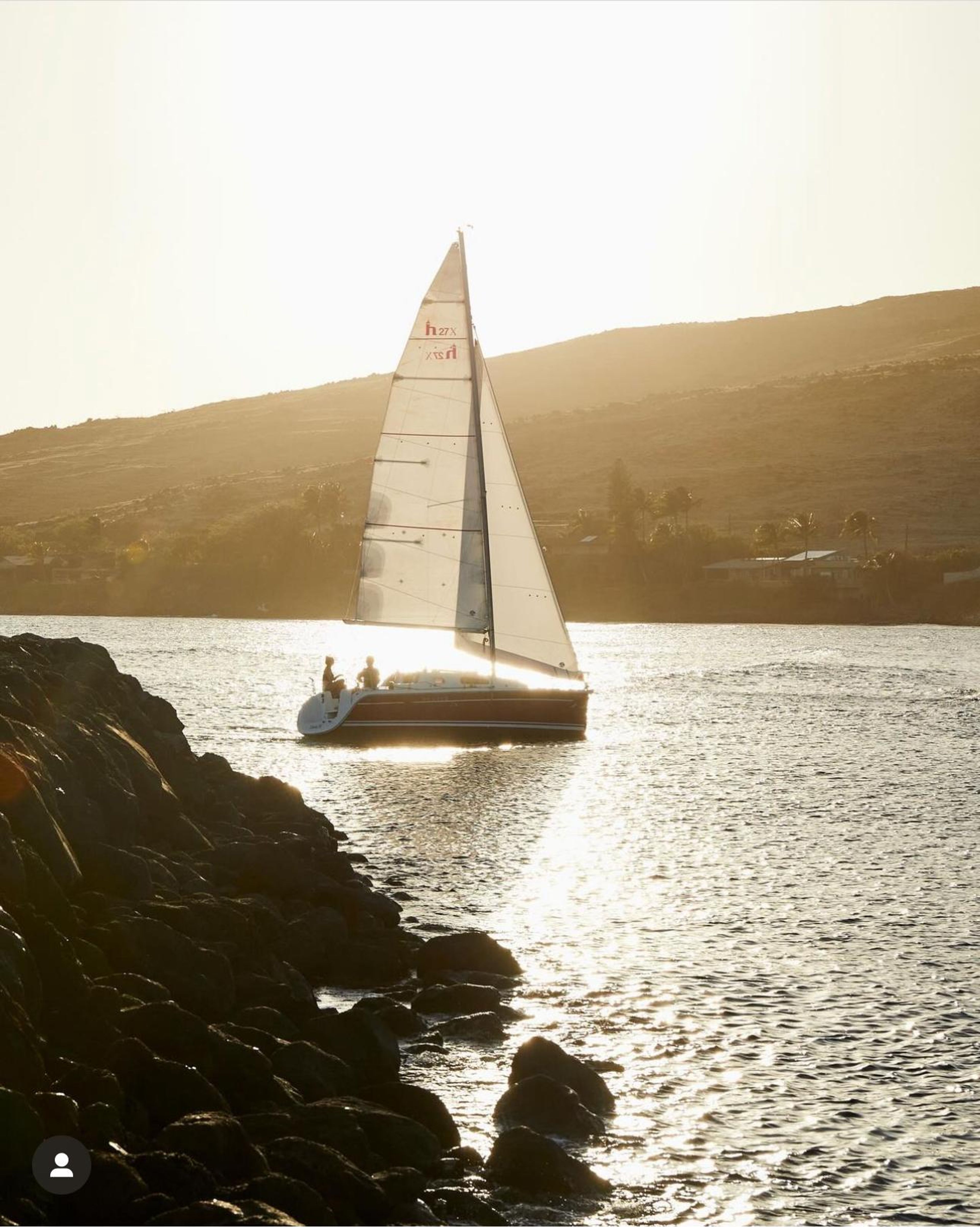 Sailboat gliding on water during sunset with rocky shoreline